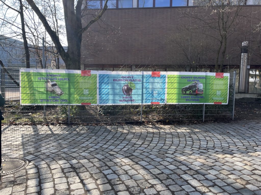 Ein Gebäude mit einem Metallzaun ist zu sehen, an dem 3 Plakate mit Umwelt-SLogans darauf schlecht zu lesen sind. 